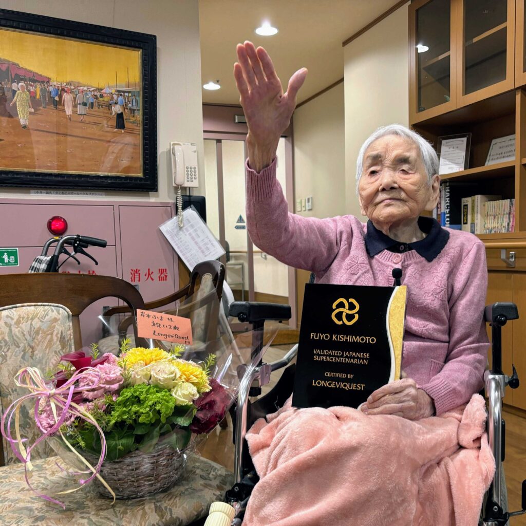 Fuyo Kishimoto raising her arm in a cheerful “Banzai” gesture, alongside a flower basket and a plaque presented by LongeviQuest to mark her verified supercentenarian status.