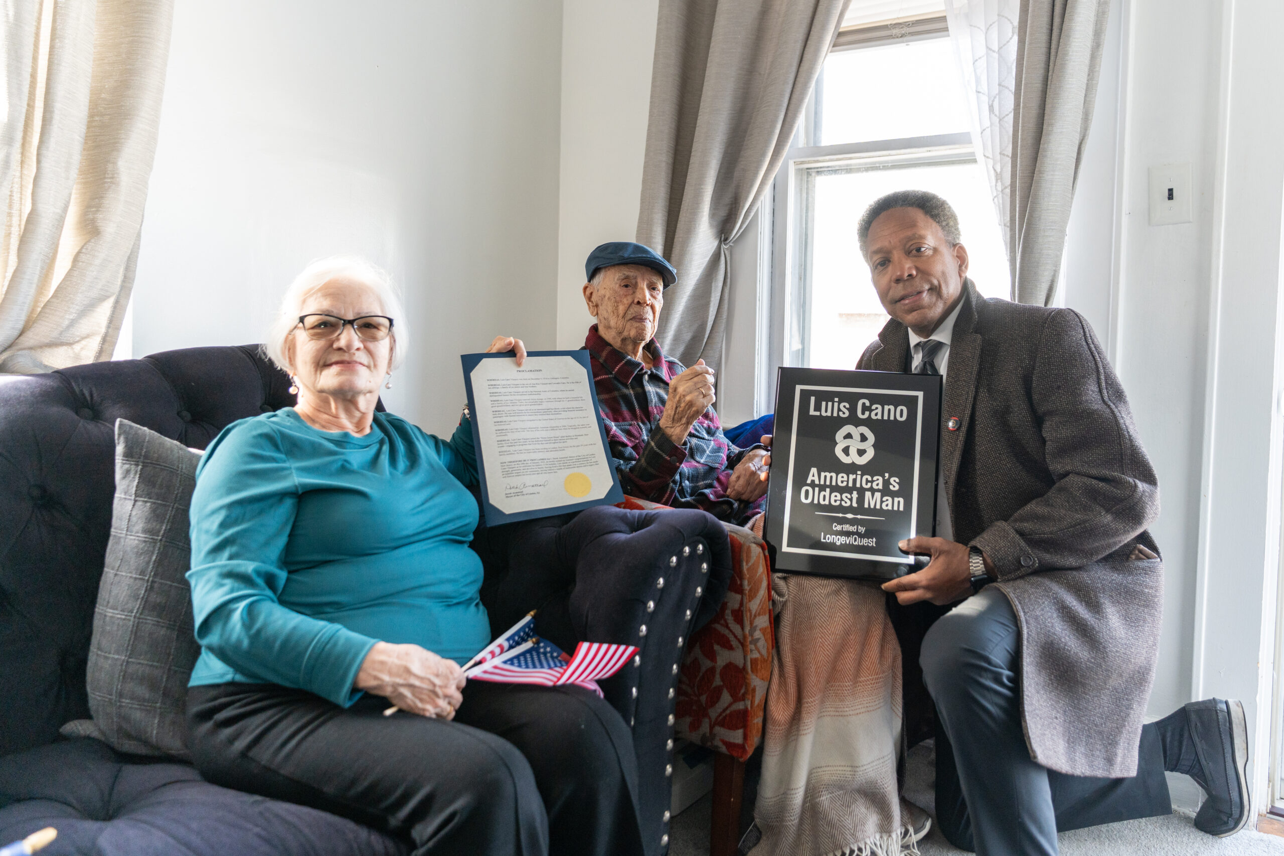 Luis Cano, America's oldest man, with his daughter Damaris and Linden, NJ Mayor Derek Armstead holding city proclamation and LongeviQuest trophy.